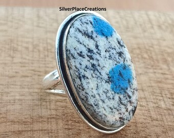 May include: Oval-shaped silver ring featuring a k2 stone with a white and gray speckled pattern and two bright blue spots. The ring has a double band design. The text "SilverPlaceCreations" is visible in the image.