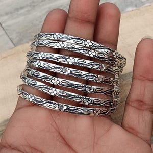 Puede incluir: Un conjunto de siete pulseras plateadas con diseños florales y de hojas. Las pulseras están apiladas una encima de la otra y se llevan en la muñeca.