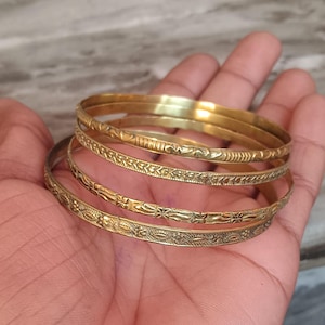 May include: Four gold-colored metal bangles with intricate engraved designs. The bangles are stacked on top of each other.