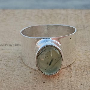 Op de afbeelding: Een brede zilveren ring met een grote, ovale, lichtgroene steen. De ring heeft een gladde, gepolijste afwerking en een dikke band. De steen heeft donkergroene insluitsels. De ring ligt op een lichtbruin houten oppervlak.