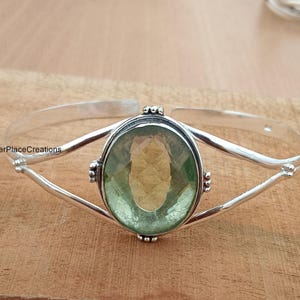 May include: A silver bangle bracelet featuring a green oval gemstone set in a silver bezel. The bracelet has a delicate, openwork design.