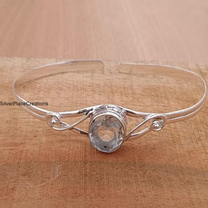 Pode incluir: Um bracelete de prata com uma pedra oval transparente. O bracelete tem um design delicado com volutas de cada lado da pedra.