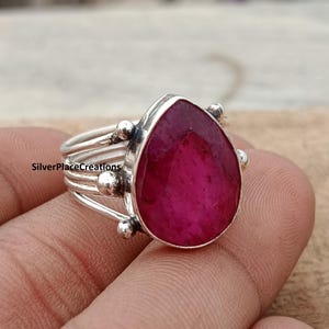 Puede incluir: Un anillo de plata con una gran gema de rubí rojo oscuro en forma de pera. El anillo tiene un diseño multibanda con pequeños detalles de cuentas de plata. El anillo se muestra en el dedo de una persona.