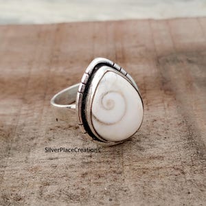 May include: A silver ring with a teardrop-shaped, white shell, displaying a spiral design. The shell is set in a silver bezel with a textured edge. The ring's band is plain, polished silver.