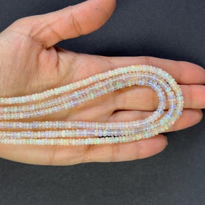 Pode incluir: Um fio de contas de opala branca, com um leve brilho iridescente, é exibido em uma mão. As contas são pequenas e redondas, e o fio é composto por várias fileiras.