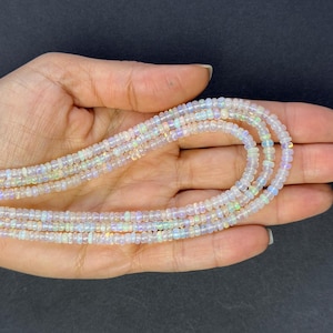 May include: A strand of white opal beads, with iridescent rainbow flashes, is displayed on a hand. The beads are small and round, and the strand is long enough to be worn as a necklace.