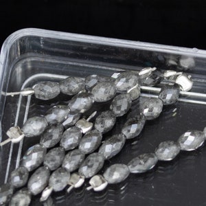 Könnte beinhalten: Eine Strähne grauer facettierter Edelsteinperlen in einem durchsichtigen Plastikbehälter. Die Perlen sind oval geformt und haben eine glatte, polierte Oberfläche.