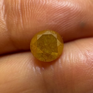 May include: A round, faceted gemstone with a vibrant golden-yellow hue. The stone features a symmetrical cut with multiple triangular and kite-shaped facets that reflect light across its surface, showcasing a translucent and warm amber-like color.