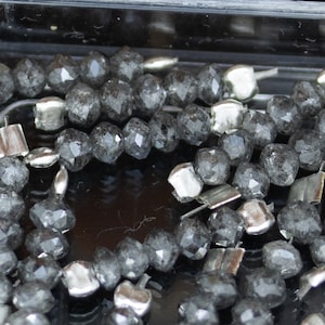 Könnte beinhalten: Nahaufnahme von facettierten, dunkelgrauen Perlen mit metallischem Glanz. Die Perlen sind mit silberfarbenen Metallteilen aufgefädelt. Die Perlen haben ein geometrisches, facettenreiches Design, das Licht reflektiert. Schmuckherstellungsbedarf.