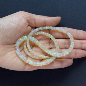 Pode incluir: Um fio de contas de opala branca com cores arco-íris iridescentes. As contas são facetadas e têm um formato ligeiramente irregular. As contas são enfiadas em um fio transparente e estão dispostas em um padrão solto e fluido.