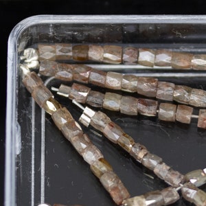 Op de afbeelding: Close-up van een streng bruine gefacetteerde edelsteen kralen in een doorzichtige plastic container. De kralen zijn rechthoekig van vorm en hebben een licht iriserende glans.