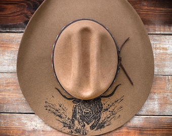 Puede incluir: Un sombrero de vaquero marrón con un gráfico de toro negro en la parte delantera. El sombrero tiene una banda de cuero marrón y una correa de barbilla de cuero. El sombrero es de fieltro.