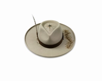 May include: A beige felt fedora hat with a brown leather band and a feather accent. The hat has a star design on the crown.