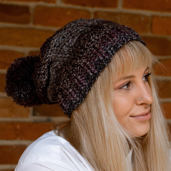 Bonnet en laine texturée multicolore avec pompon amovible et long revers. Tons marron, gris et bordeaux. Le brassard peut être porté plié ou à plat.