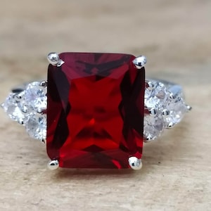 May include: A silver ring with a large, red, square-shaped gemstone surrounded by smaller, clear gemstones.