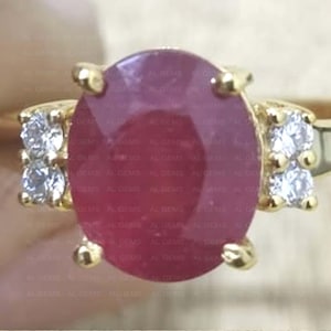 Peut inclure: Une bague en or ornée d'un grand rubis ovale rouge foncé. De petites pierres précieuses claires sont serties de chaque côté du rubis. La bague présente un design classique et élégant.