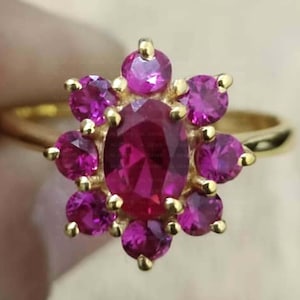 Può includere: Un anello d'oro con un rubino centrale ovale circondato da rubini rotondi più piccoli, che formano un motivo floreale. La fascia dell'anello è in oro liscio. I rubini sono di un rosa intenso. Un gioiello.