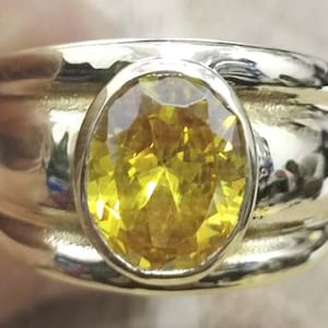 Peut inclure: Une bague dorée ornée d'une grande pierre précieuse jaune de forme ovale. La bague a un large anneau avec un motif texturé. La pierre précieuse est le point central, avec une couleur jaune vibrante et des facettes complexes.