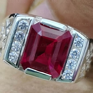 May include: A silver ring featuring a large, square-shaped, red gemstone surrounded by smaller, clear gemstones.