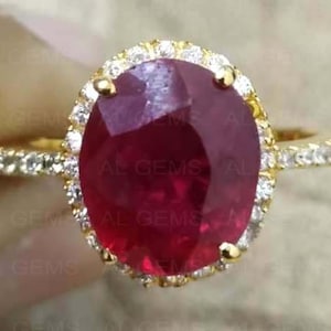 Può includere: Un anello d'oro con un grande rubino ovale. Il rubino è circondato da un alone di piccole pietre preziose trasparenti. La fascia dell'anello è anch'essa ornata da pietre preziose più piccole, creando un effetto scintillante.