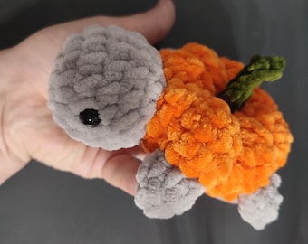 May include: A handmade plush turtle with a pumpkin design. The turtle has a gray head and feet, an orange body, and a green stem. The plush toy is made of soft, textured material and features a black eye.