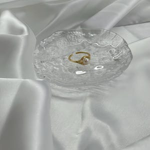 Op de afbeelding: Een goudkleurige, met draad omwikkelde ring met een enkele, ronde, witte parel. De ring ligt op een heldere, getextureerde glazen schaal, tegen een achtergrond van witte, vloeiende stof.