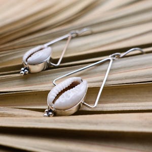 May include: A pair of silver-toned earrings featuring white cowrie shells. The shells are set in a silver metal frame with a triangular design. The earrings have a small cluster of decorative beads at the base of the shell.