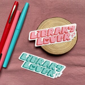 Puede incluir: Dos pegatinas con el texto "Library Lover" en una fuente retro. La pegatina superior es rosa con un corazón rosa y brillos. La pegatina inferior es verde azulado con un corazón verde azulado y brillos.