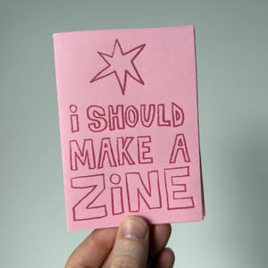 Pode incluir: Um pequeno zine rosa com a frase "I SHOULD MAKE A ZINE" e uma estrela. O zine é segurado por uma mão. O texto é em estilo manuscrito. O zine é um pequeno livreto, possivelmente feito à mão.