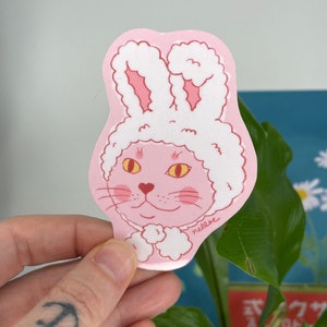 May include: Pink cat wearing a white fluffy bunny costume with pink ears. The cat has yellow eyes and a heart-shaped nose. The sticker has the word "nellose" written on the bottom.