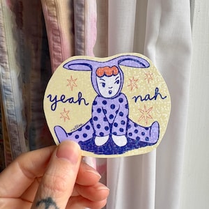 Peut inclure: Un autocollant jaune avec un bébé dessin animé portant une combinaison à pois violets et blancs et des oreilles de lapin. Le bébé est assis les jambes croisées et a une expression surprise sur son visage. L'autocollant dit "yeah nah" en texte orange et blanc.