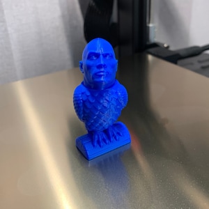 May include: A blue 3D printed figurine of a person with a bald head and a serious expression. The figurine is wearing a textured, scaly outfit and is standing on a small base.