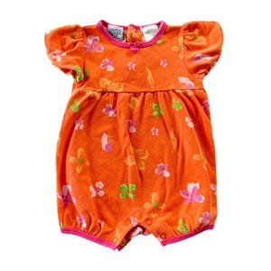 May include: An orange baby romper with a floral pattern. The romper features short sleeves and a round neckline with pink trim. The floral pattern includes pink, orange, and green flowers. The romper has snap closures at the bottom.
