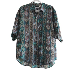 Puede incluir: Una camisa de manga corta con botones y un estampado geométrico en tonos turquesa, marrón, gris y rosa. La camisa tiene cuello y un bolsillo en el pecho. La camisa está colgada en una percha blanca.