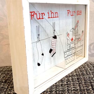 Könnte beinhalten: Ein weißer Holzrahmen mit den roten Worten "Für ihn" und "Für sie". Das Kunstwerk zeigt Socken, die an einer Wäscheleine hängen, und Schuhe auf Kisten.