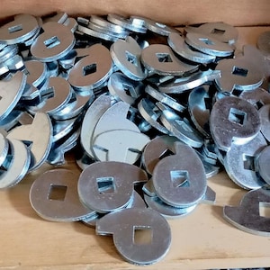 Könnte beinhalten: Ein Haufen silberfarbener Metallscheiben mit quadratischem Loch in der Mitte und einer gebogenen Kerbe an einer Seite. Die Scheiben liegen auf einer Holzoberfläche.