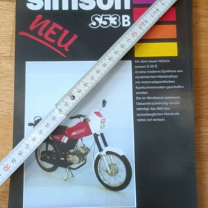 Könnte beinhalten: Eine schwarze Werbung für ein Simson S53B Moped mit dem Wort "NEU" in Rot. Ein weißes und rotes Moped ist abgebildet. Ein Lineal liegt diagonal über der Werbung und misst etwa 30 cm.