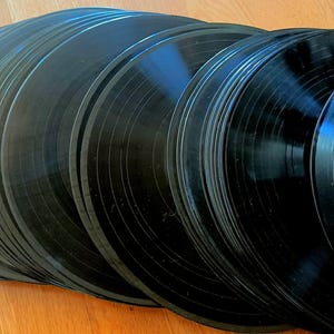 Puede incluir: Una colección de discos de vinilo negros, apilados y abanicados, con surcos visibles. Los discos están dispuestos sobre una superficie de madera marrón claro, creando un contraste entre los discos oscuros y el fondo más claro. La imagen resalta la textura y el brillo del vinilo.