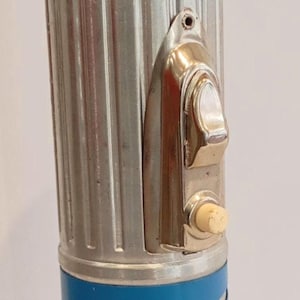 Può includere: Torcia vintage con base e parte superiore blu, e corpo argentato e scanalato. La torcia ha un interruttore cromato e un piccolo pulsante rotondo. Design retrò classico.