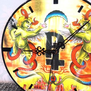 May include: A round wall clock with a unique, colorful design. The clock face features two green figures with horns and wings, surrounded by orange and yellow hues. The clock hands are black, and the overall theme is artistic and unconventional.