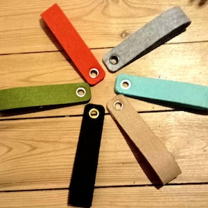 May include: A collection of six felt keychains in various colors: red, gray, teal, tan, black, and green. Each keychain has a metal grommet for attaching keys. The keychains are arranged in a circular pattern on a wooden surface.