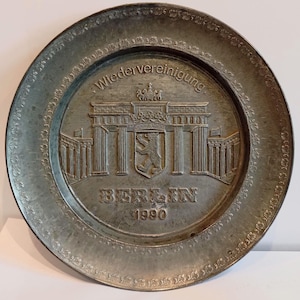Könnte beinhalten: Eine silberfarbene Zierplatte mit dem Brandenburger Tor, einem Wappen und dem Text "BERLIN 1990". Die Platte hat einen detaillierten Rand und das Wort "Wiedervereinigung" steht oben.