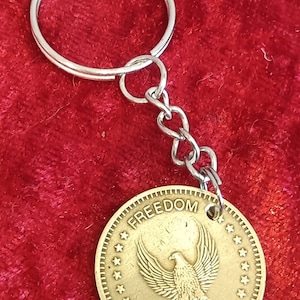 Könnte beinhalten: Ein goldfarbener Schlüsselanhänger mit einem runden Anhänger mit einem Adler und dem Wort „FREEDOM“. Der Anhänger ist an einer silbernen Kette und einem Ring befestigt. Der Hintergrund ist ein roter Stoff.