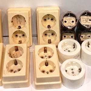 Può includere: Una collezione di prese e spine elettriche vintage di varie forme e dimensioni. Le prese sono principalmente bianco sporco e beige, con alcuni componenti neri e marroni. L'assortimento comprende prese doppie e singole, insieme a diversi tipi di spine.