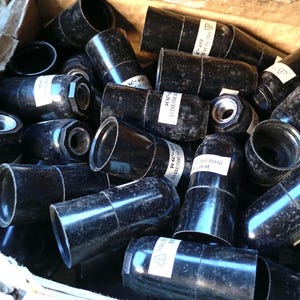 May include: A pile of black, cylindrical metal components with a shiny surface. Some have white labels with text and numbers. The objects have a flared end and a threaded interior. The components are in a cardboard box.