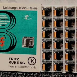 Peut inclure: Une boîte de relais électriques avec une étiquette turquoise et blanche. L'étiquette contient du texte en allemand et un schéma. Les relais sont dans un plateau en plastique blanc. Chaque relais a des composants noirs et cuivrés.