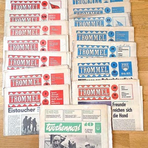 May include: A collection of vintage newspapers titled "Trommel" and "Wochenpost" are displayed. The newspapers feature red and blue text, with headlines and articles in German. The image includes various issues, some with dates and issue numbers visible.