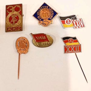 May include: A collection of vintage pins and badges, featuring red, gold, and black colors. Some pins display flags, while others have text like "DDR" and "1961". The pins vary in shape, including rectangles, flags, and round designs.