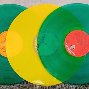Puede incluir: Tres discos de vinilo en tonos verdes y amarillos. Los discos tienen etiquetas naranjas y rojas. Los discos están dispuestos en un patrón semicircular, con el disco amarillo en el centro.