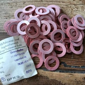 Könnte beinhalten: Ein Haufen rosa Gummischeiben mit einem weißen Papieretikett. Das Etikett hat schwarzen Text, darunter "VEB Fahrzeug-und Jagdwallenwerk Ernst Thälmann Suhl" und Zahlen. Die Scheiben liegen auf einer Holzoberfläche.